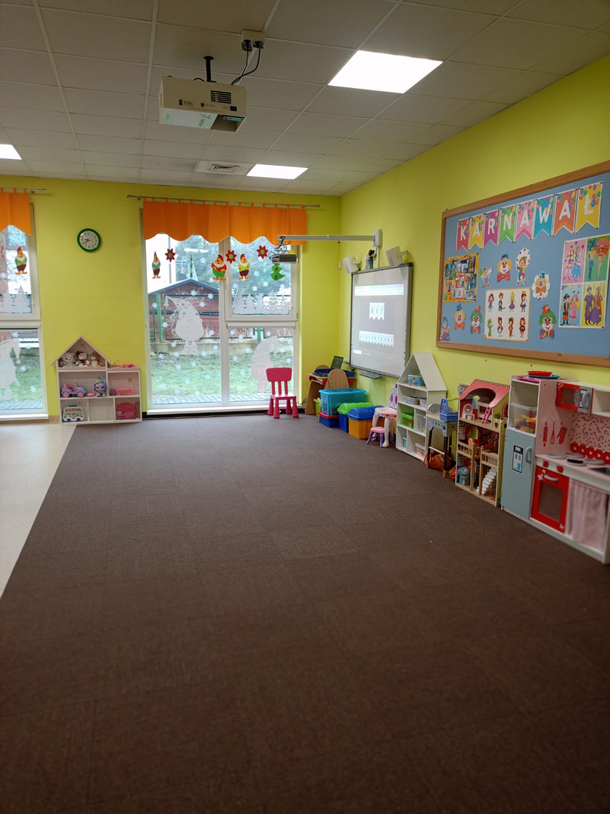 Zdjęcie ukazuje przestronną i funkcjonalną salę przedszkolną, która została wyposażona z myślą o edukacji i zabawie dzieci. Jasne ściany, duże okna i przemyślane rozmieszczenie elementów wyposażenia sprawiają, że pomieszczenie jest przyjazne i dostosowane do potrzeb najmłodszych. Wnętrze zaprasza do aktywnego spędzania czasu i rozwijania umiejętności dzieci.  Na ścianie po prawej stronie zamontowano nowoczesną tablicę interaktywną, która umożliwia prowadzenie ciekawych i angażujących zajęć edukacyjnych.   Duże okna– zapewniają naturalne światło, a jednocześnie ozdobione są wesołymi dekoracjami świątecznymi, które wprowadzają wyjątkowy nastrój   Wydzielony kącik zabaw – po prawej stronie znajdują się różnorodne zabawki i akcesoria, w tym kuchnia zabawkowa, które rozwijają wyobraźnię i umiejętności społeczne dzieci.  Miękka wykładzina– pokrywająca dużą część podłogi, idealna do bezpiecznej zabawy oraz ćwiczeń ruchowych.